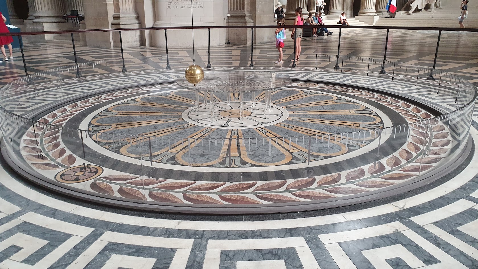 O pêndulo de Foucault no Panthéon, em Paris: a esfera de latão suspensa sobre um grande disco de mármore com escala graduada e pinos dispostos ao longo da circunferência, cercado por um guarda-corpo, com visitantes ao fundo.